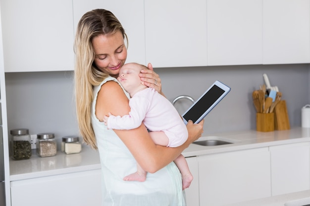 Madre usando tableta digital mientras carga a su bebé en la cocina