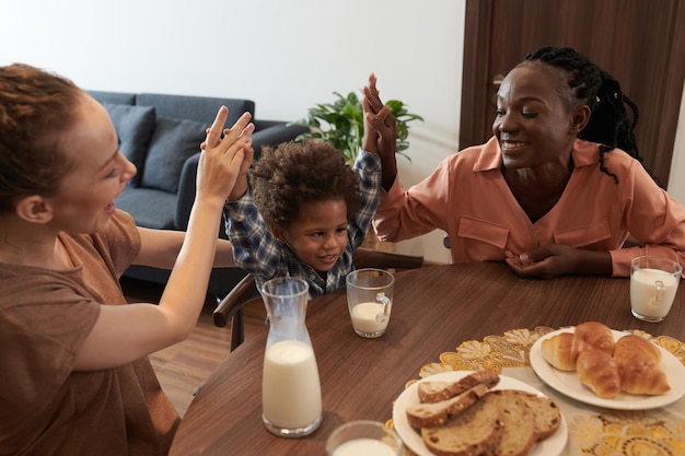 Madres felices dando cinco a un hijo pequeño sentado en la mesa del desayuno
