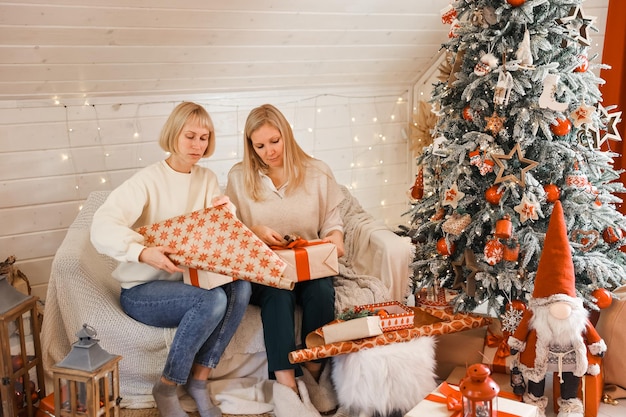 Las madres felices de las mujeres se están preparando para la Navidad, los regalos del paquete de año nuevo en casa mientras están sentados cerca del árbol de Navidad. Vacaciones de invierno, concepto de decoración del hogar