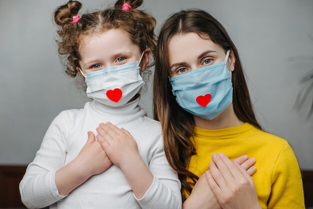 Las madres y la hija que se toman de las manos en el pecho se sientan en la cama, usan una máscara facial con el corazón como una forma de mostrar agradecimiento a los médicos y enfermeras por la ayuda en la lucha contra la enfermedad. COVID-19