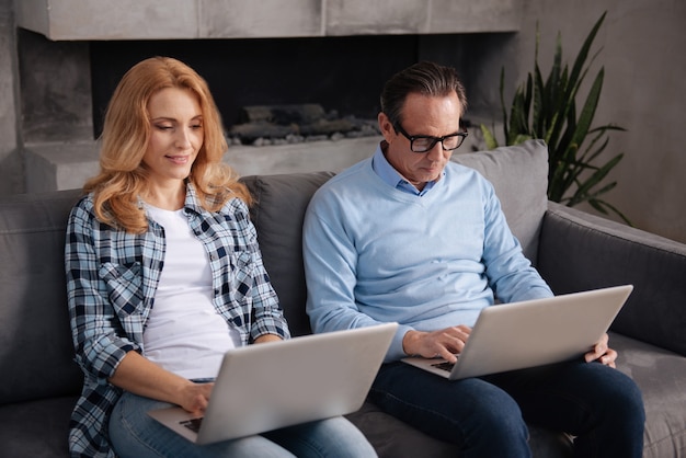 Madura mujer encantadora positiva sentada en casa y usando computadoras portátiles con padre anciano mientras navega por Internet y envía mensajes de texto