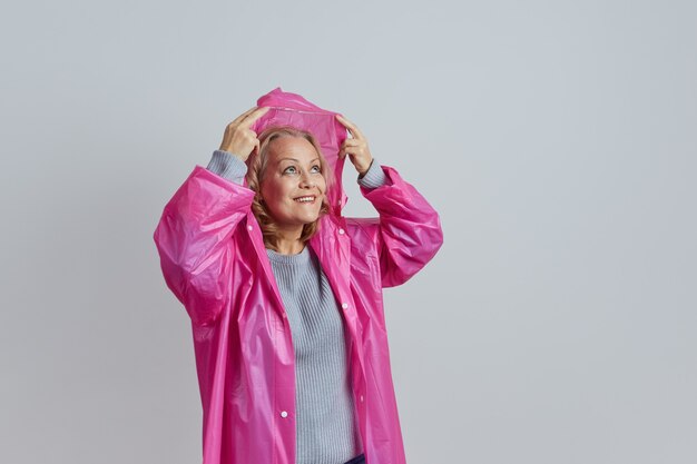 Madura rubia sonriente con un impermeable magenta se esconde bajo una capucha de la lluvia.