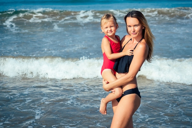 Mãe abraçando a filha em pé de maiô na praia