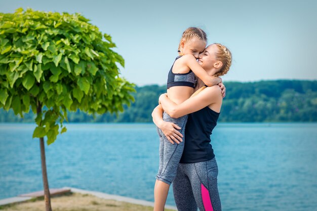 Mãe abraçando a filha perto do lago vestindo roupas esportivas ao ar livre