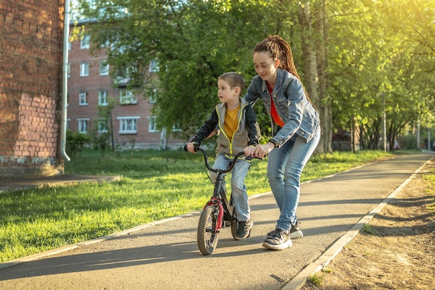 Mãe ajuda um menino a aprender a andar de bicicleta de duas rodas no parque Uma agradável férias de esportes de verão para crianças