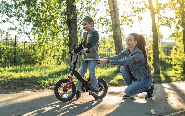 Mãe ajuda um menino a aprender a andar de bicicleta de duas rodas no parque Uma agradável férias de esportes de verão para crianças