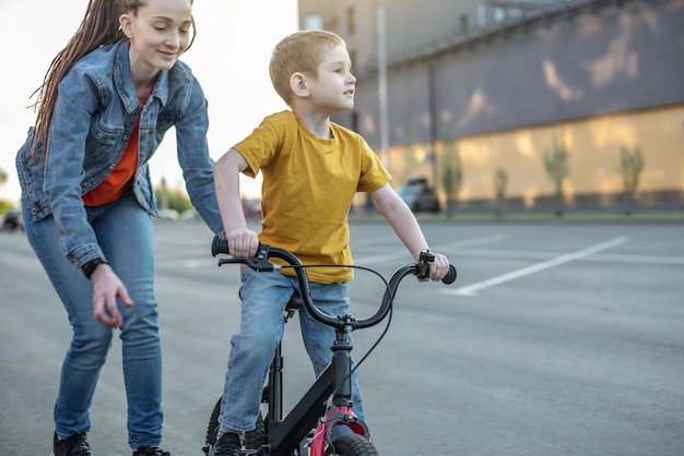 Mãe ajuda um menino a aprender a andar de bicicleta de duas rodas no parque uma agradável férias de esportes de verão para crianças