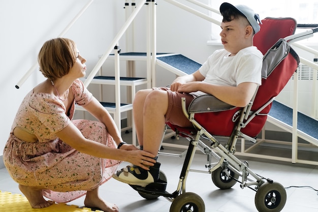 Mãe ajudando menino deficiente em cadeira de rodas com paralisia cerebral a calçar sapatos