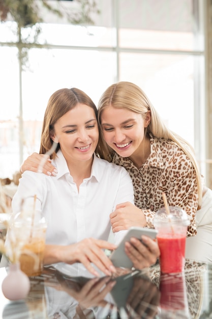 Mãe alegre com smartphone fazendo selfie com sua filha carinhosa enquanto ambas passam um tempo no café após as compras