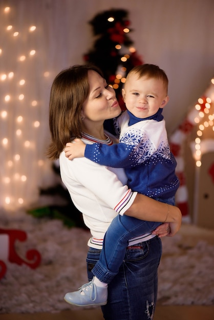 Mãe alegre e seu filho lindo filho se divertindo perto da árvore dentro de casa. Família amorosa na sala de Natal.