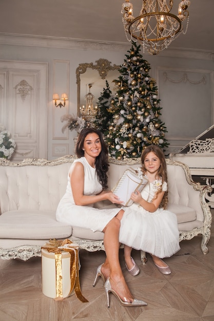 Mãe alegre e sua linda filha trocando presentes no interior clássico branco no contexto de um piano e uma árvore de Natal decorada.