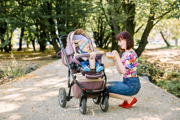 Mãe alimentando seu bebê com uma colher. Mãe dando comida para criança de dez meses andando com carrinho de bebê no parque. Comida de bêbe.