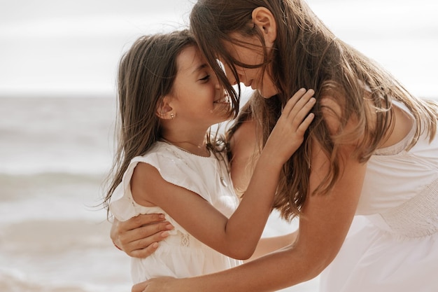 Mãe amorosa em vestido branco abraçando criança feliz enquanto passa tempo na praia em fundo desfocado do mar ondulante no verão