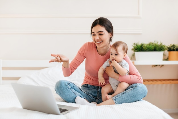 Mãe animada segurando bebê e fazendo videochamada com laptop sentado na cama e gesticulando para