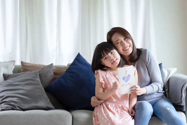 Mãe asiática abraça sua filha fofa que dá um cartão feito à mão com a palavra "Eu amo mamãe" para surpreendê-la em casa