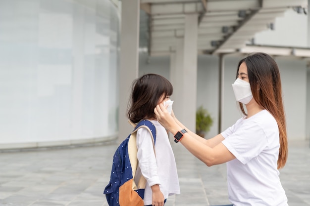 Mãe asiática ajuda sua filha usando uma máscara médica para proteção contra Covid-19 ou surto de coronavírus a se preparar para ir à escola quando voltar à escola.