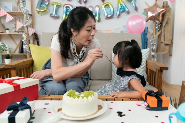 mãe asiática carinhosa alimentando bolo de creme de filha bebê fofo com uma colher enquanto comemora o aniversário juntos em casa com decorações coloridas