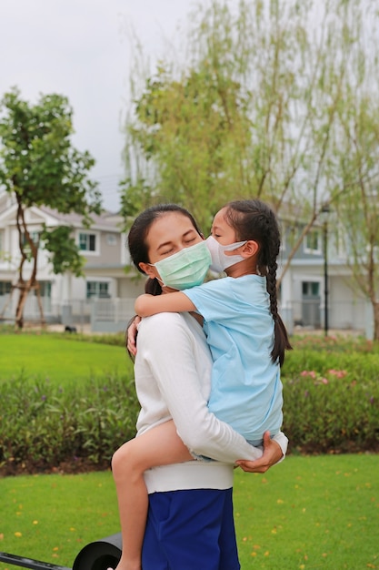 Mãe asiática carregando a filha com máscara protetora no rosto e se beijando em um jardim público durante o surto de coronavírus e gripe