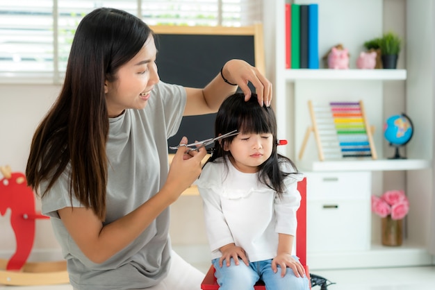 Mãe asiática cortando cabelo para a filha na sala de estar em casa enquanto fica em casa a salvo do Covid-19 Coronavirus durante o bloqueio. Auto-quarentena e conceito de distanciamento social.