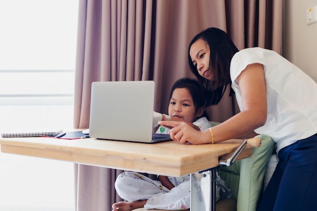 Mãe asiática ensinando sua filha doente, aprendendo on-line no computador laptop, enquanto está no hospital. Ensino doméstico de isolamento e ensino à distância.