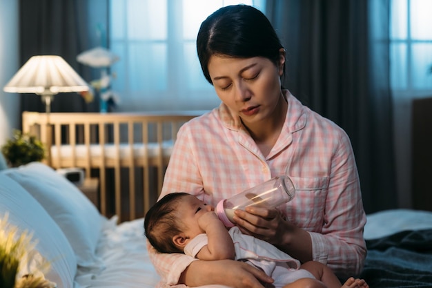 mãe asiática exausta está dormindo cochilando enquanto está sentado à beira da cama dando leite engarrafado para seu bebê tarde da noite em um quarto escuro com pouca luz em casa.