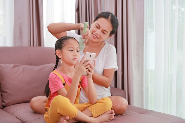 Mãe asiática penteando o cabelo da filha enquanto brinca de smartphone no sofá da sala de casa