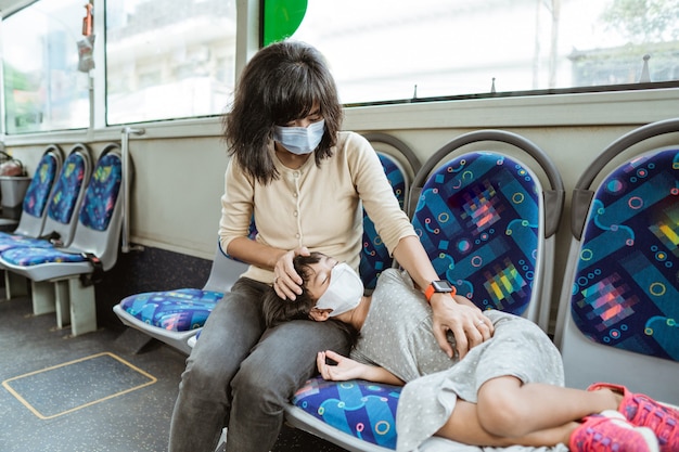 Mãe asiática usando máscara junto com uma filha que dorme em um banco durante uma viagem de ônibus