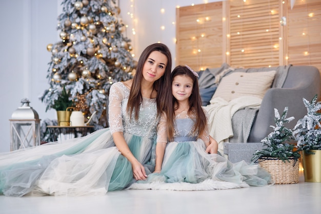 Mãe atraente e sua filha em um suave vestidos de noite, sentada no chão perto da árvore de Natal em uma sala aconchegante.