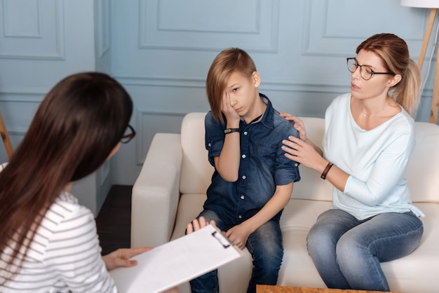 Mãe atraente usando roupas casuais sentada perto do filho no sofá enquanto o acalma