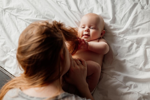 Mãe beija os pés descalços de um bebê recém-nascido sorridente deitado na cama jovem com um bebê em casa