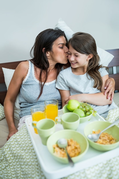 Mãe beijando nas bochechas da filha enquanto tomando café da manhã na cama