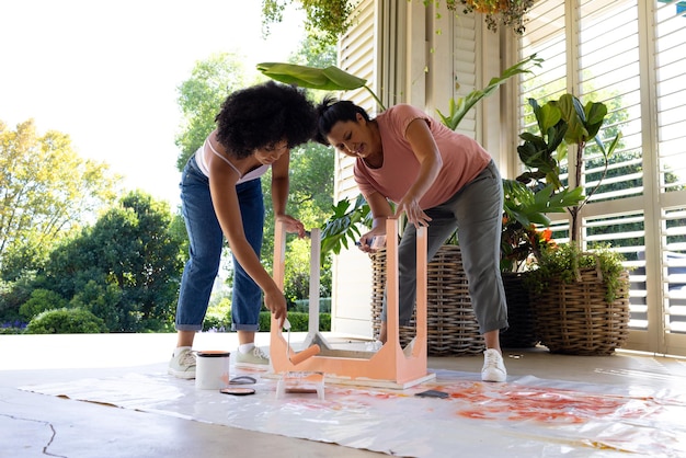 Foto mãe biracial e filha adulta estão pintando uma cadeira de rosa fora de casa como um projeto de upcycling