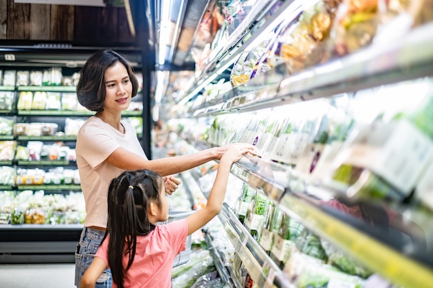 Mãe bonita asiática nova que guarda a cesta do mantimento com sua criança que anda no supermercado.