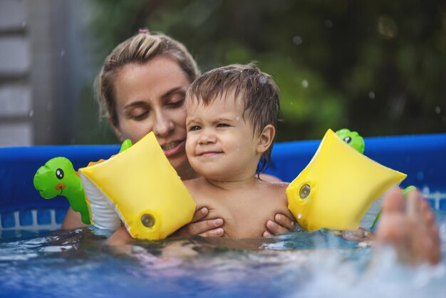 Mãe brinca com um bebê nu em mangas na piscina no contexto de um pôr do sol de verão