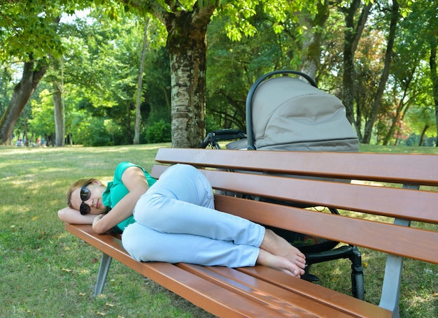 Mãe cansada dormindo em um banco em um parque com um bebê em um carrinho