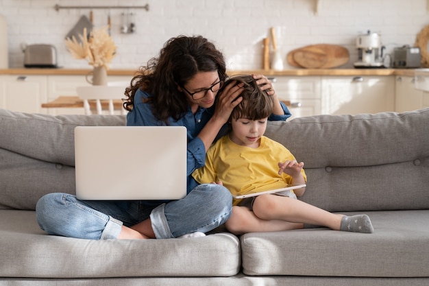 Mãe carinhosa ajuda criança pré-escolar com e-learning no tablet digital enquanto trabalha no laptop de casa