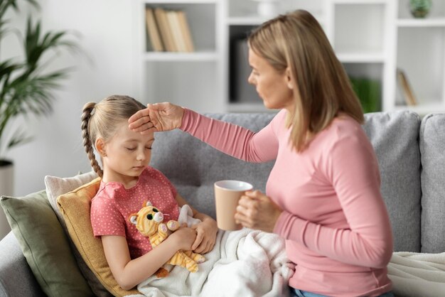 Mãe carinhosa dando chá para filhinha doente e tocando sua testa