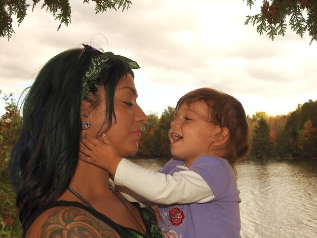 Foto mãe carregando filha bonita enquanto está de pé ao lado do lago contra o céu durante o pôr do sol