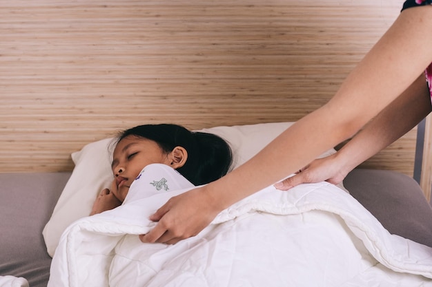 Foto mãe colocando um cobertor para a filha na cama criança asiática dormindo no quarto