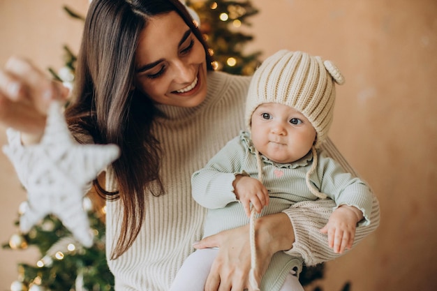 Mãe com a filha segurando um brinquedo de natal perto da árvore de natal