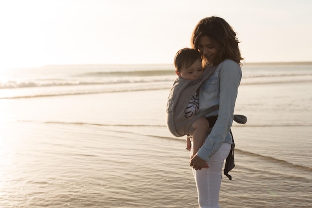 Mãe com ergobaby carregando criança na praia