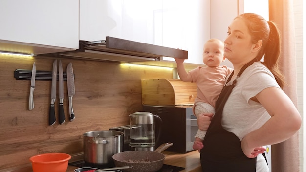 Mãe com expressão cansada cozinha e cuida de menina