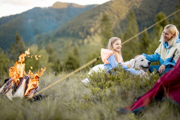 Mãe com filha e cachorro sentam perto da fogueira perto da tenda na natureza