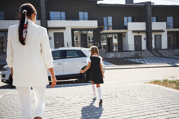 Mãe com filha em uniforme escolar ao ar livre perto de carro branco.