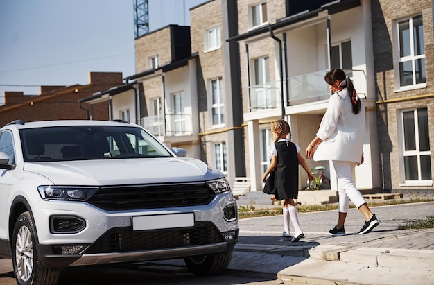 Mãe com filha em uniforme escolar ao ar livre perto de carro branco.