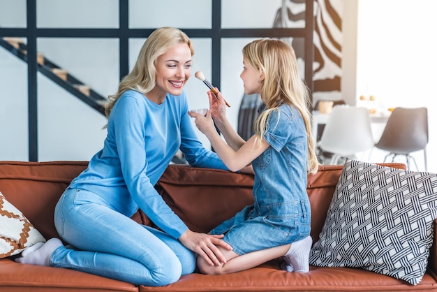 Mãe com filha se divertindo e fazendo maquiagem cosmética no sofá em casa