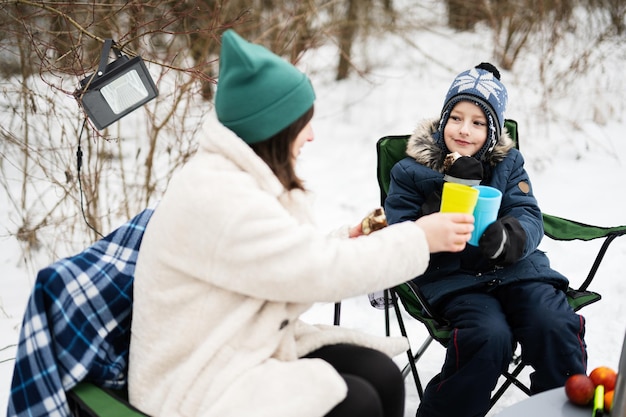 Mãe com filho brinda com chá quente enquanto fazem piquenique na floresta de inverno