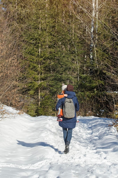 Mãe com filho nos braços e mochileiro caminhando pela estrada coberta de neve no contexto da floresta de coníferas Dia de inverno Moldura vertical