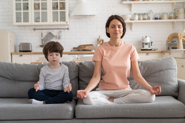 Mãe com filho pequeno fazendo exercícios de ioga sentados juntos no sofá em casa ensinando a criança a meditar