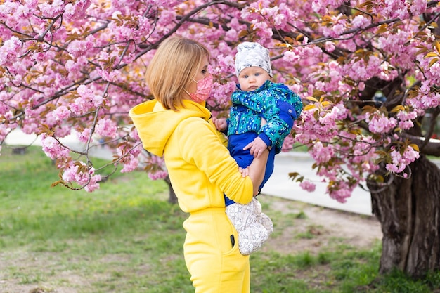 Mãe com máscara médica e filho pequeno na natureza brincando no parque primavera Garotinho e mãe se divertem na atividade de fim de semana nos jardins floridos de Sakura